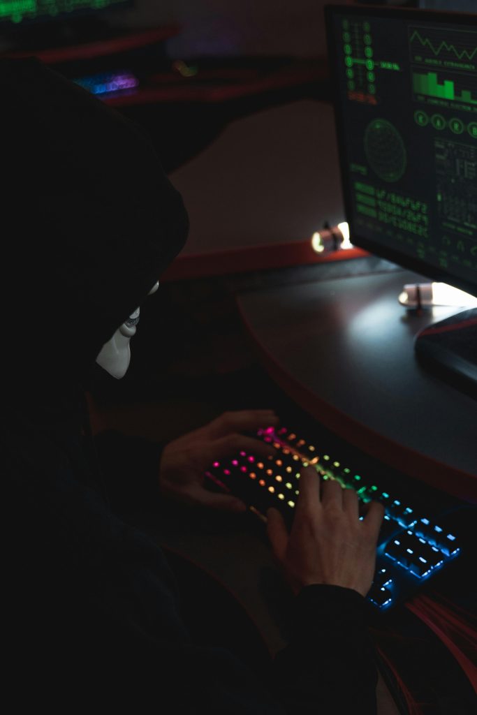 An anonymous hacker in a black hoodie typing on a colorful RGB keyboard, highlighting cybersecurity themes.