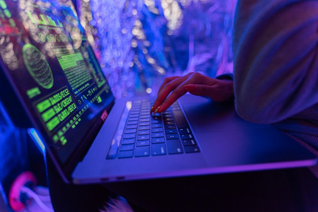 zipwp-image-5475752 Close-up of hands typing on a laptop displaying cybersecurity graphics, illuminated by purple light.